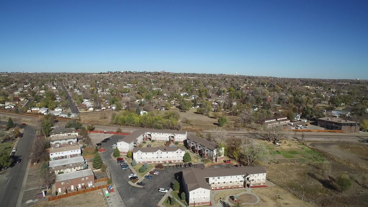 Drone-height view in Westminster, Colorado with low-rise residential buildings in the foreground, a straight road through the neighborhood, dry fields, and no visible people.