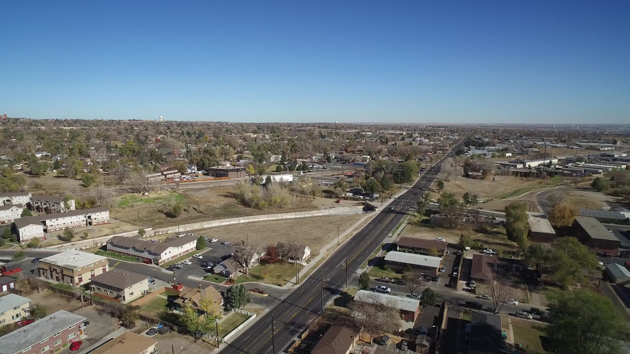This Westminster, Colorado aerial frame keeps the road near center while showing business rooftops, parking areas, residential edges, sparse trees, and no visible people.