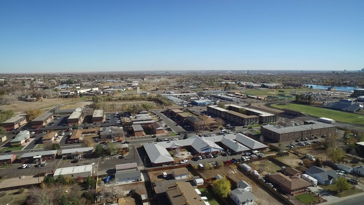 Wide aerial view over suburban houses, roads, commercial buildings, green fields, lake, and distant skyline.