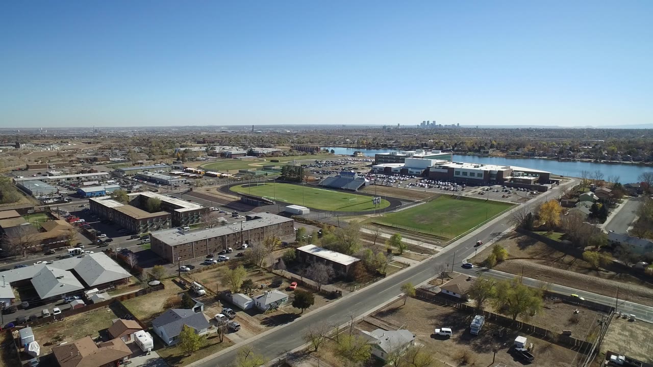 The Westminster, Colorado aerial view centers on the athletic field and nearby buildings, with a road crossing the foreground and the lake stretching behind them, no visible people.