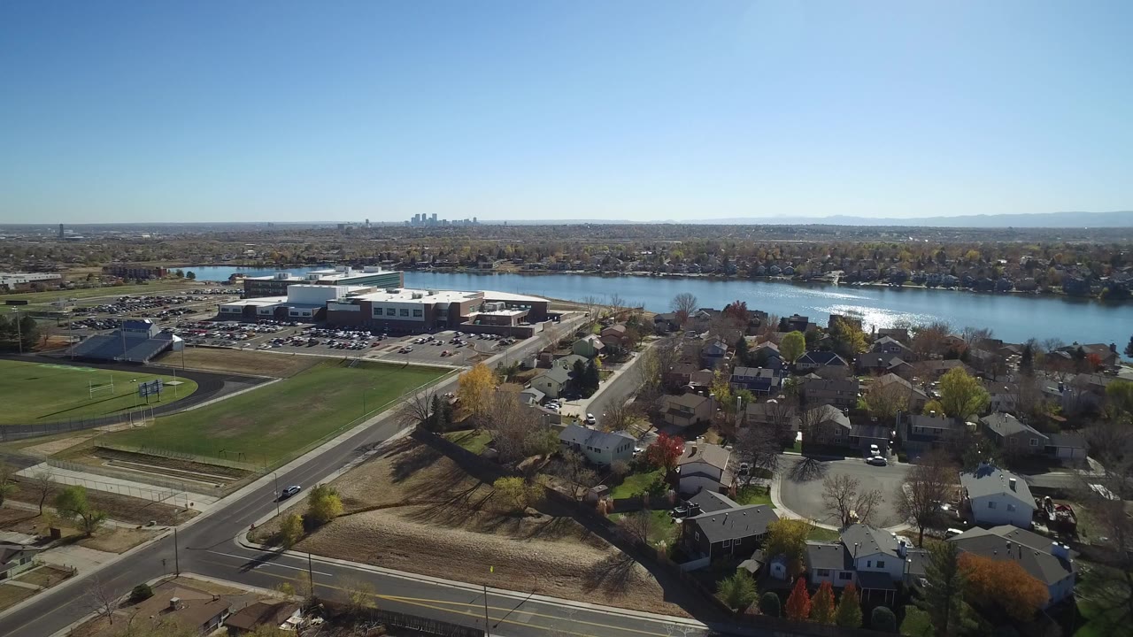 High aerial view over Westminster, Colorado homes and roads toward a bright band of water, athletic fields, a large building, distant skyline, and no visible people.