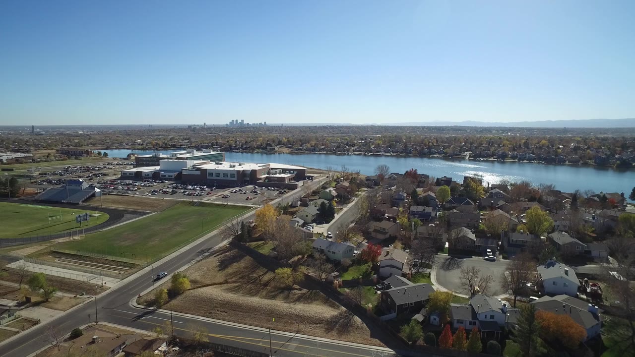 Aerial frame in Westminster, Colorado looking across residential roofs and a green field toward a large low building, reflective water, distant skyline, and no visible people.