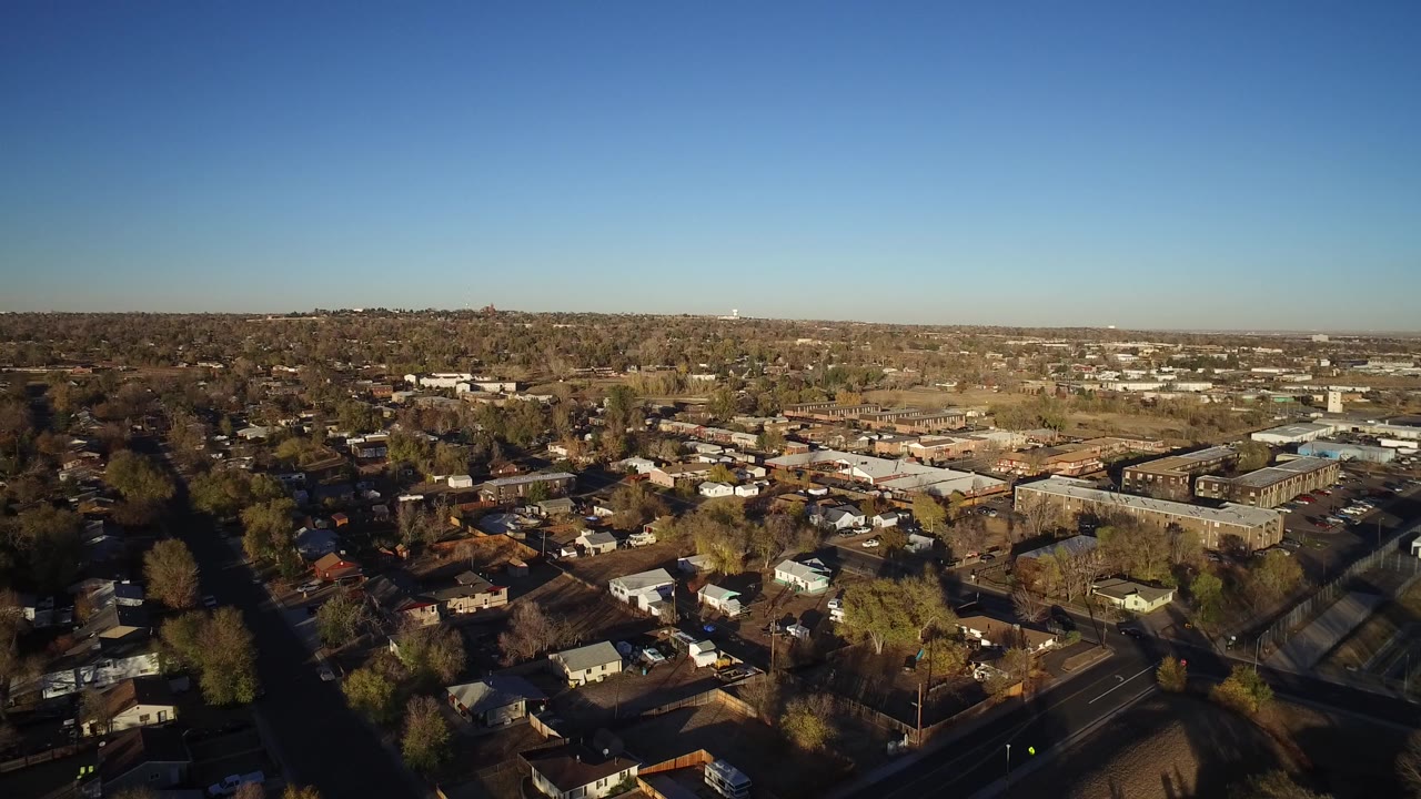 Aerial view over suburban houses, trees, roads, and low buildings under a clear sky.