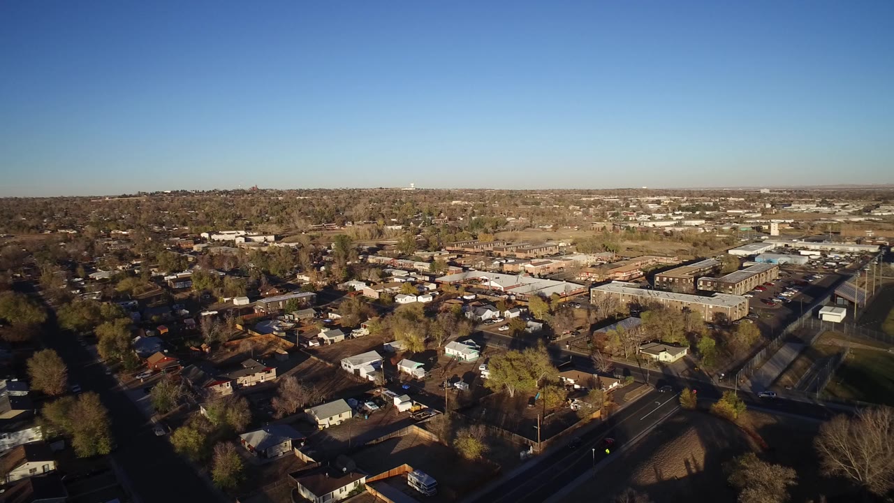 Aerial view of suburban homes, streets, trees, and distant low-rise buildings under blue sky.