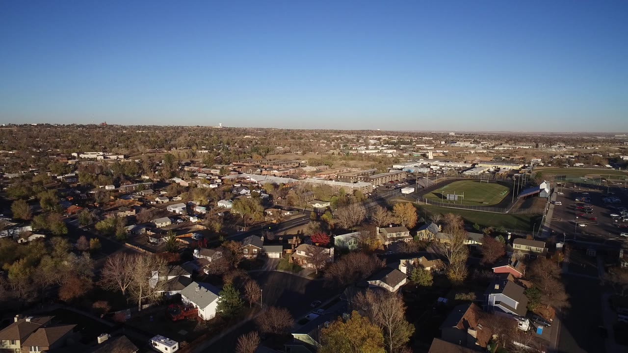 Aerial view of suburban houses, trees, streets, and a sports field under blue sky.