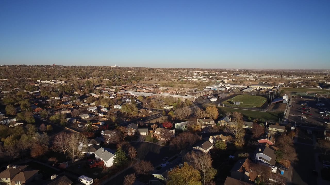Aerial frame over Westminster, Colorado showing dense rooftops, bare and colored trees, a green athletic field, parking lots, and no visible people.