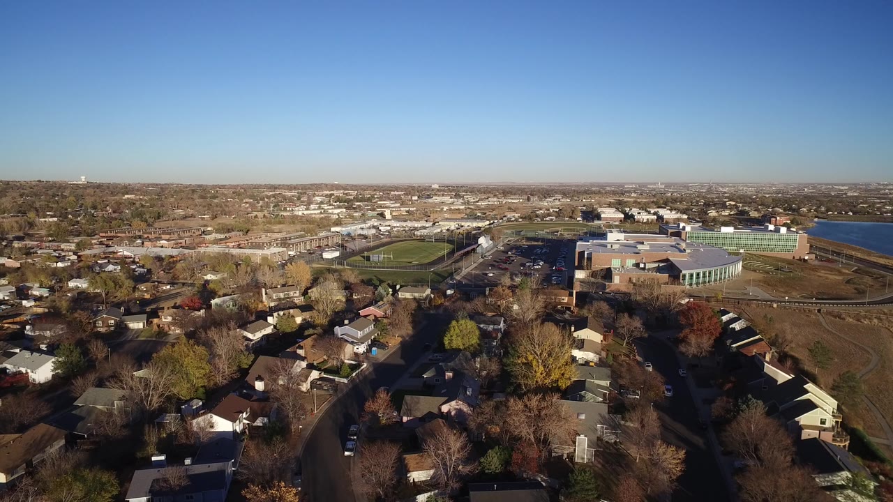 Aerial view of suburban houses, trees, athletic field, and lake edge under clear sky.