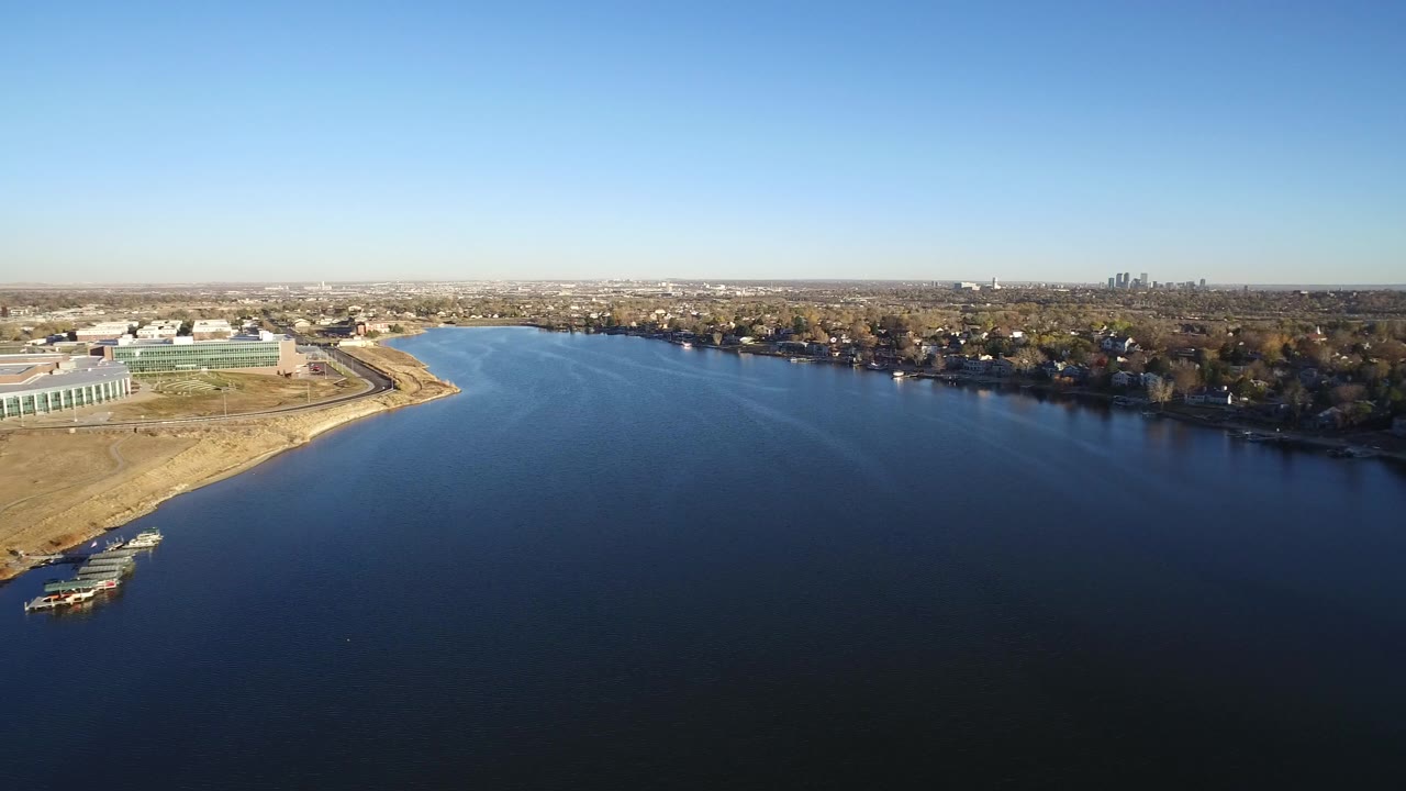 Opening aerial frame over Westminster, Colorado water with low buildings at left, houses and trees along the far shore, distant towers on the horizon, and no visible people.