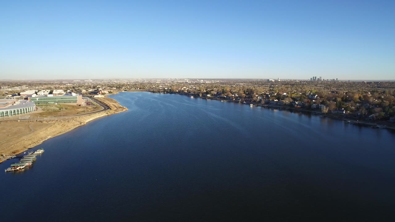 Second Westminster aerial frame shows more open blue water in front, pale shoreline land and a low green-roofed building at left, distant houses and trees, and no visible people.