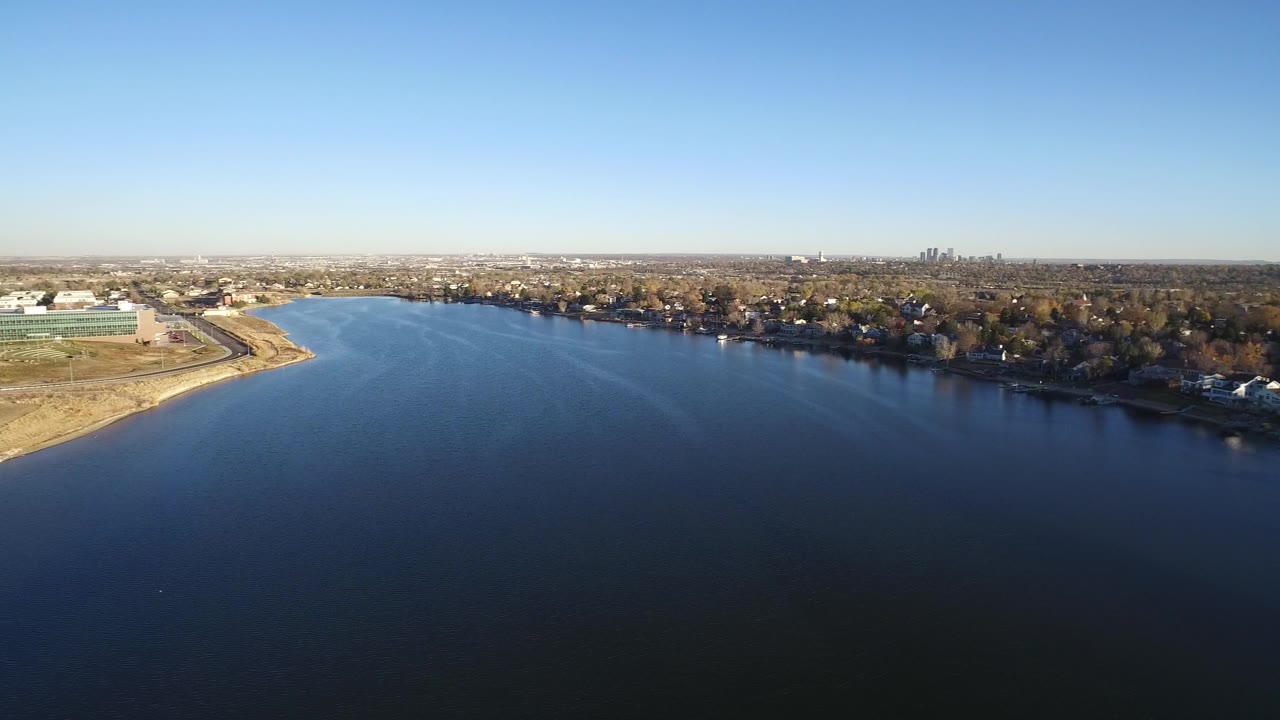 Aerial frame of open water with a residential tree line and distant skyline under clear sky.