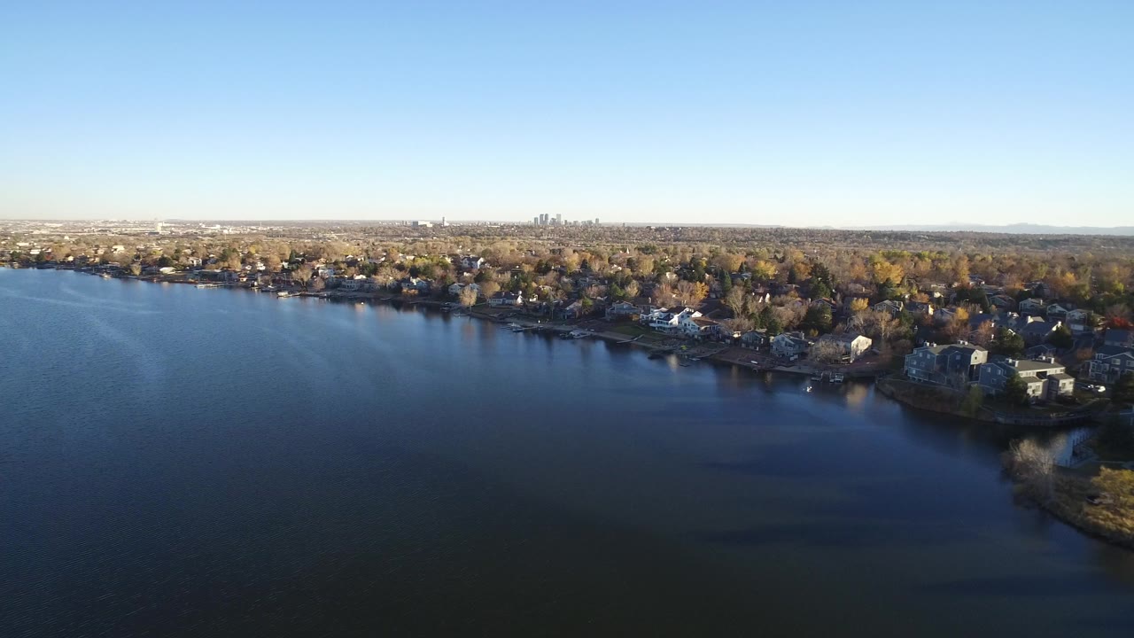 Wide aerial view in Westminster, Colorado, with dark lake water in the foreground, shoreline homes and trees beyond it, a distant city skyline on the horizon, and no visible people.