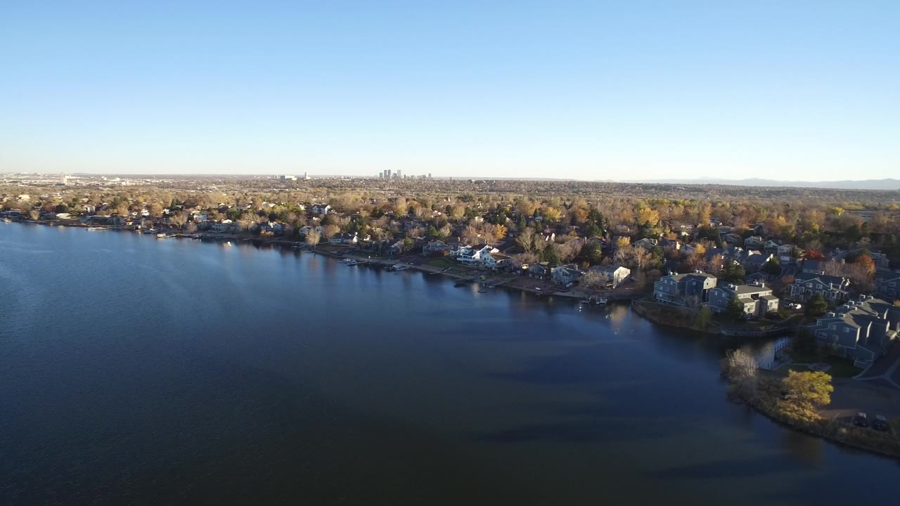 Drone-height view of a lake, residential shoreline, trees, and far city buildings.