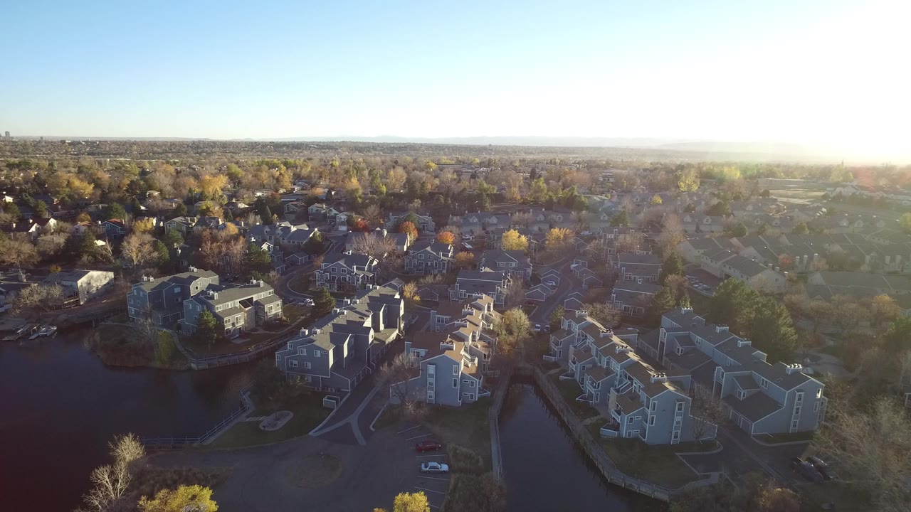 High aerial view over a Westminster, Colorado apartment complex with a dark pond at lower left, pale buildings in the foreground, autumn trees beyond, bright sun glare at right, and no visible people.