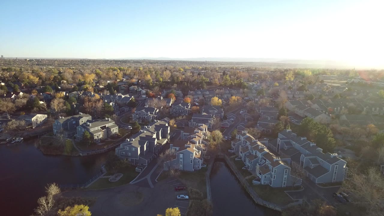 Aerial frame over Westminster, Colorado apartments with two water areas at the lower edge, curved internal roads, rows of nearby houses, distant horizon, sun haze to the right, and no visible people.