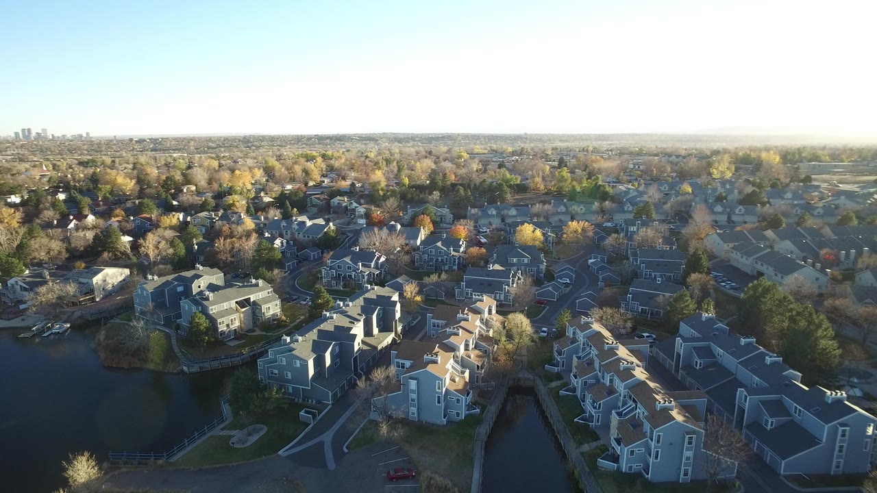 Opening aerial frame over Westminster, Colorado, showing lakeside homes, tree-lined neighborhoods, pale sky, distant skyline, and no visible people.