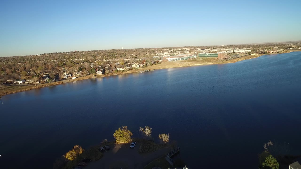 Westminster, Colorado aerial frame with the lake-like water filling most of the image, a band of homes and autumn-colored trees along the opposite shore, distant buildings, and no visible people.