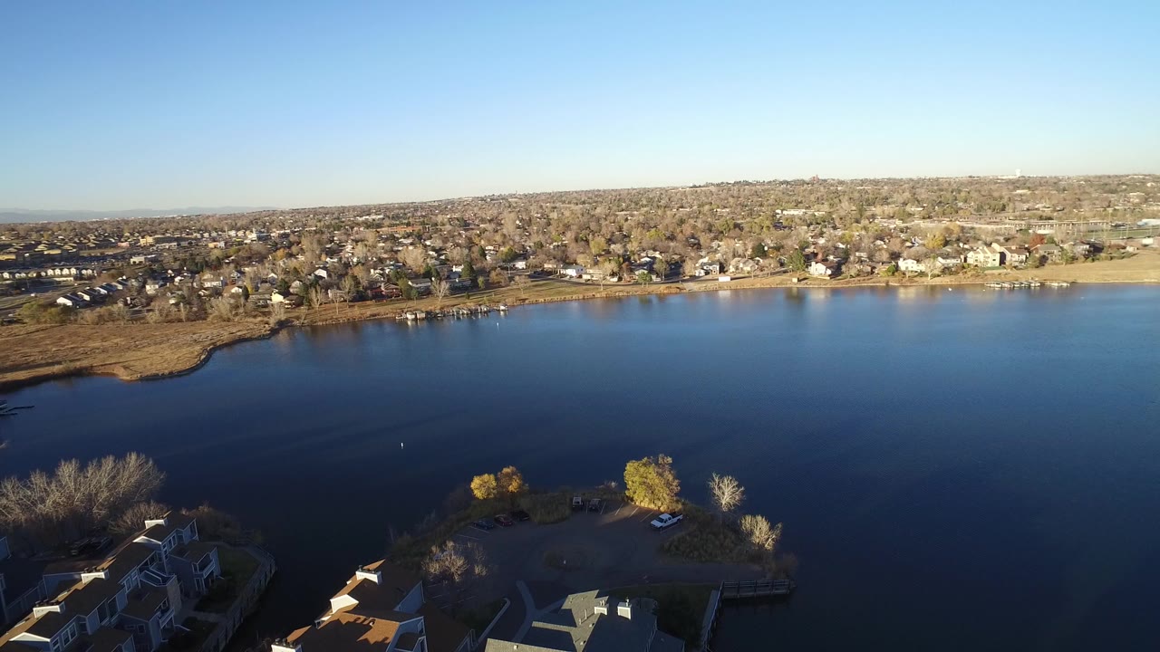 Aerial view in Westminster, Colorado looking over a dark blue lake, with townhome roofs at the bottom edge, houses and trees across the far shore, and no visible people.