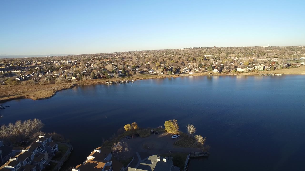 A slightly shifted aerial frame over Westminster, Colorado shows the lake filling the middle of the image, residential streets and houses beyond the far bank, and no visible people.