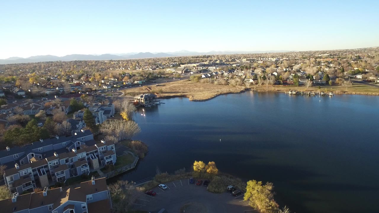 Aerial view over Westminster, Colorado with a dark lake centered between foreground apartments, surrounding neighborhoods, distant mountains, and no visible people.