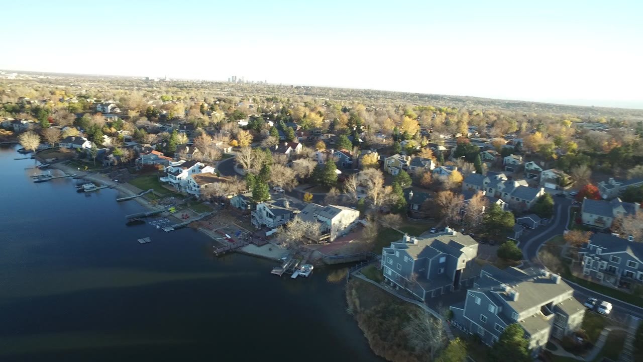 The second frame keeps the lake on the left and centers large shoreline houses, docks, curving roads, autumn tree cover, pale blue sky, and no visible people in Westminster, Colorado.