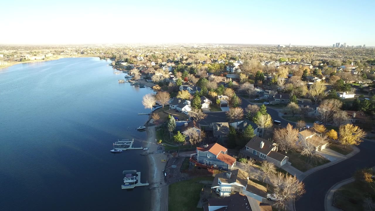 Aerial frame in Westminster, Colorado with the lake filling the left side, several docks near the shore, houses among bare and evergreen trees, and no visible people.