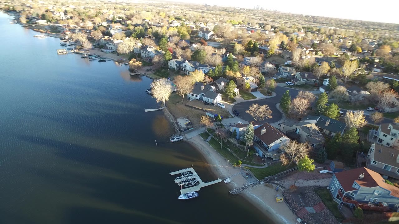 The aerial angle holds over Westminster lakefront houses, showing a curved shoreline, moored boats, residential streets, and long shadows across the water, with no visible people.