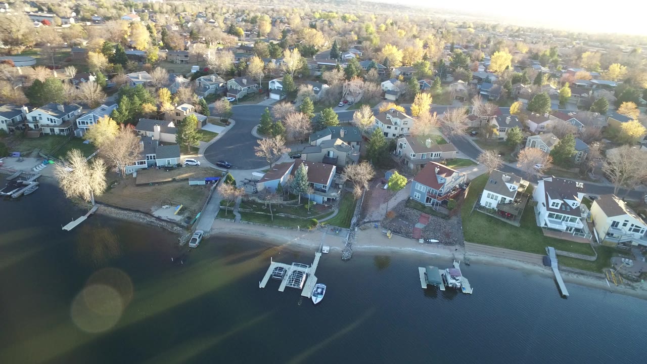 Aerial view over Westminster, Colorado lakefront homes with docks and boats along the shore, curved residential streets beyond, bright late afternoon haze, and no visible people.