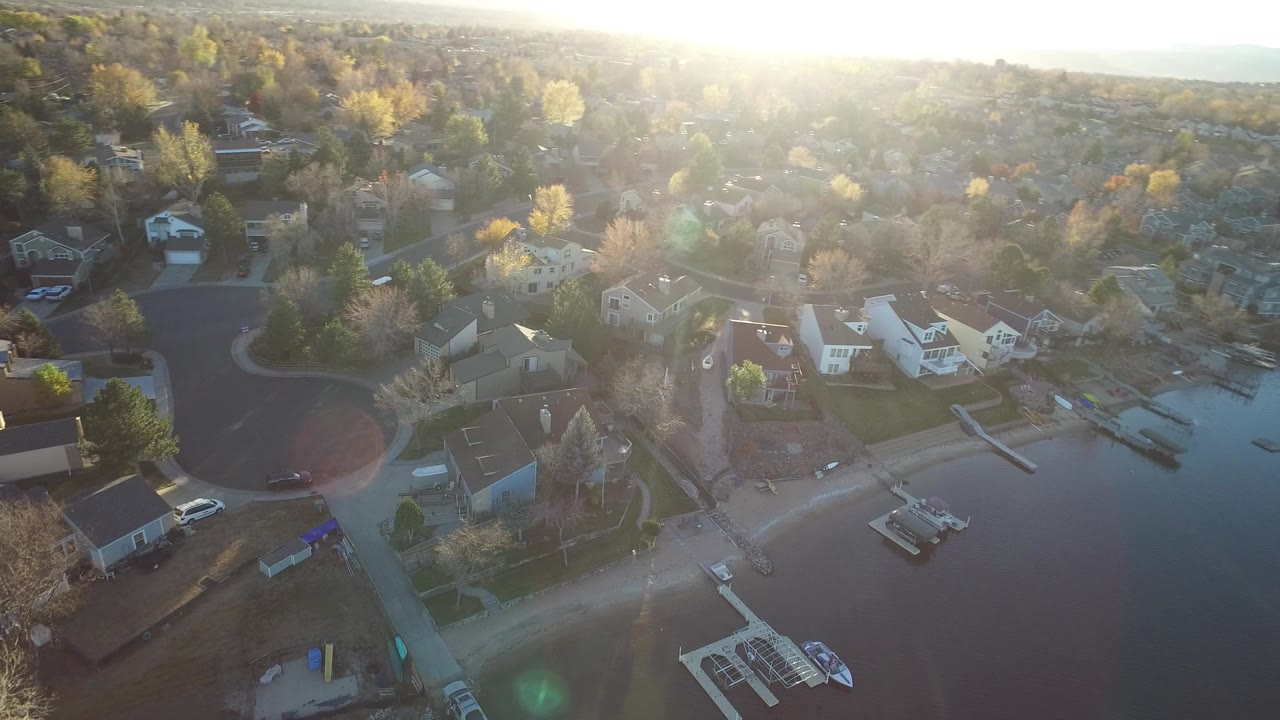 Aerial frame over Westminster, Colorado showing lakeside homes, private docks, leafless and yellow trees, bright sun glare, and no visible people.