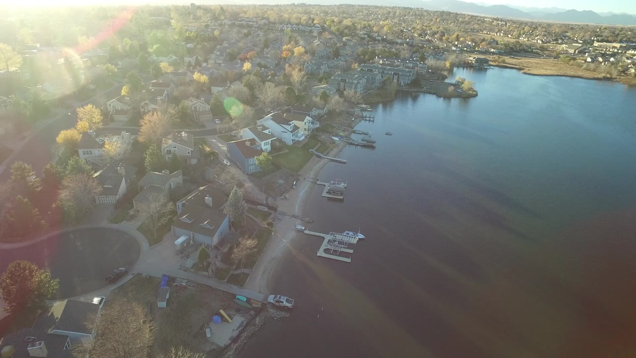 Westminster, Colorado aerial view with houses and yards in the lower frame, small docks along the shoreline, blue-green lake water to the right, lens flare, and no visible people.
