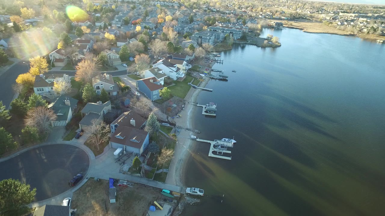 Aerial view over Westminster, Colorado lakefront homes with small docks, moored boats, a curved street, sun flare, and no visible people.