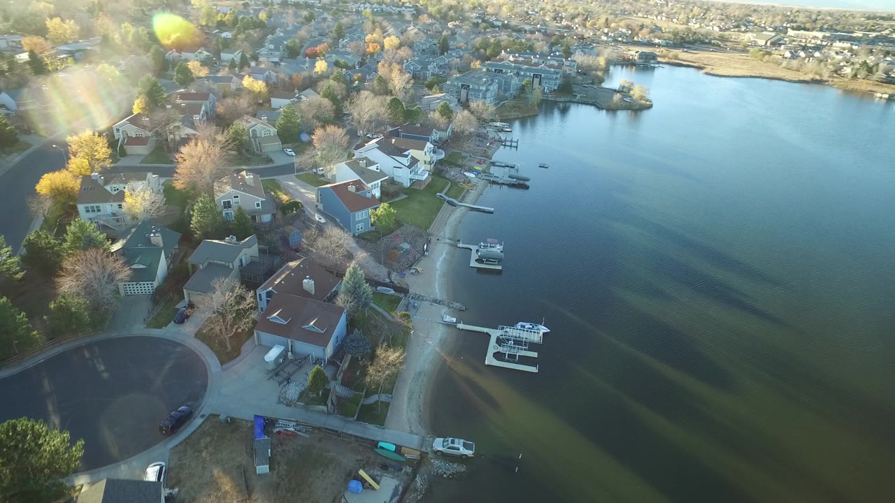 High aerial frame in Westminster, Colorado showing houses along the left shoreline, docks extending into blue-green water, distant homes across the lake, and no visible people.