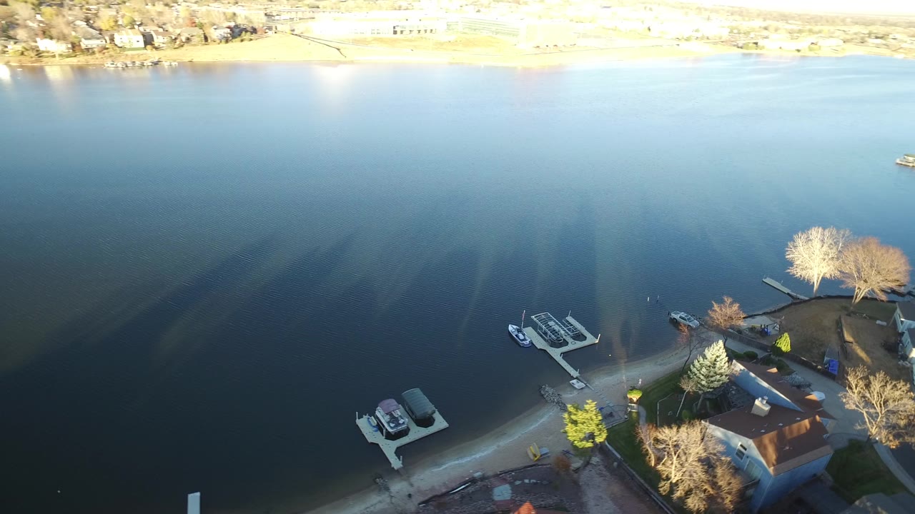 Aerial frame in Westminster, Colorado, showing calm blue water, two small dock areas, boats near shore, leafless trees, and residential roofs along the bottom edge.