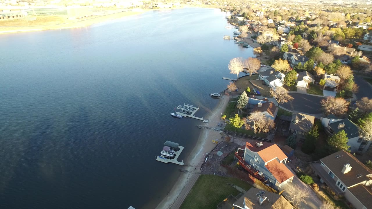 Aerial view in Westminster, Colorado with lake water filling the left side, homes and docks along the shore, small boats near the bank, and no visible people.