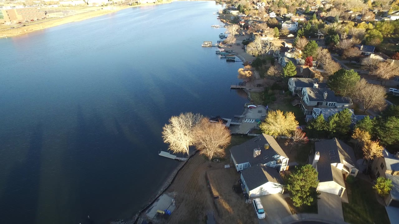 Aerial frame over Westminster, Colorado with still water filling most of the left side, lakeside houses and leafless trees along the right, and no visible people.