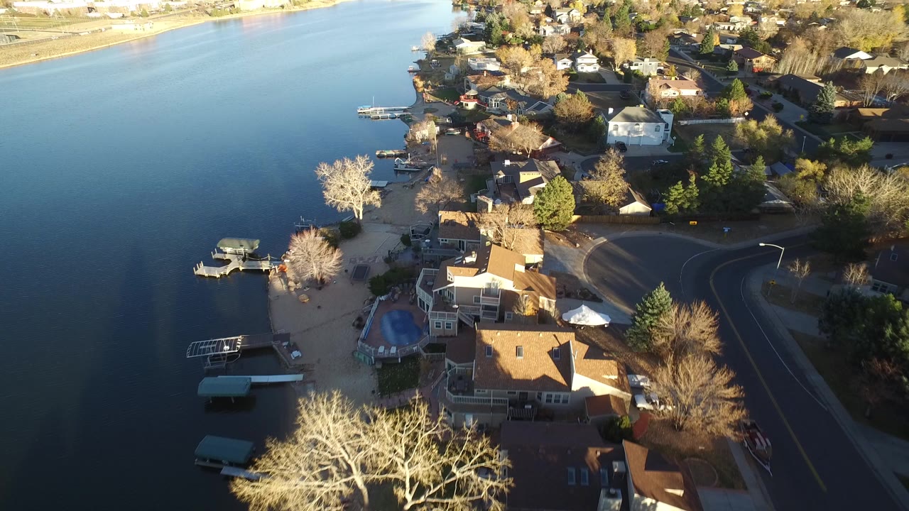 Aerial view over Westminster, Colorado homes along calm blue water, with docks, bare trees, a curved road, and no visible people.