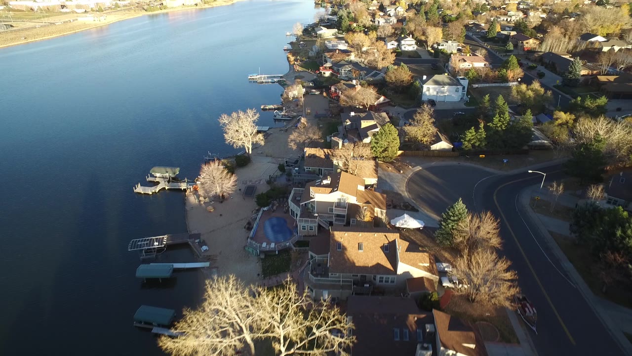 Elevated view of Westminster, Colorado shoreline homes, small docks, leafless trees, and a residential road beside still water, with no visible people.
