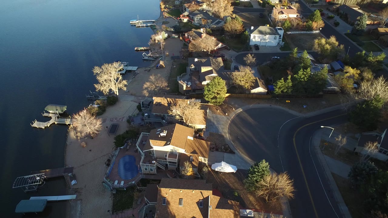 High aerial view over Westminster, Colorado houses beside a lake, with docks along the left edge, a curving road on the right, warm sunlight, and no visible people.