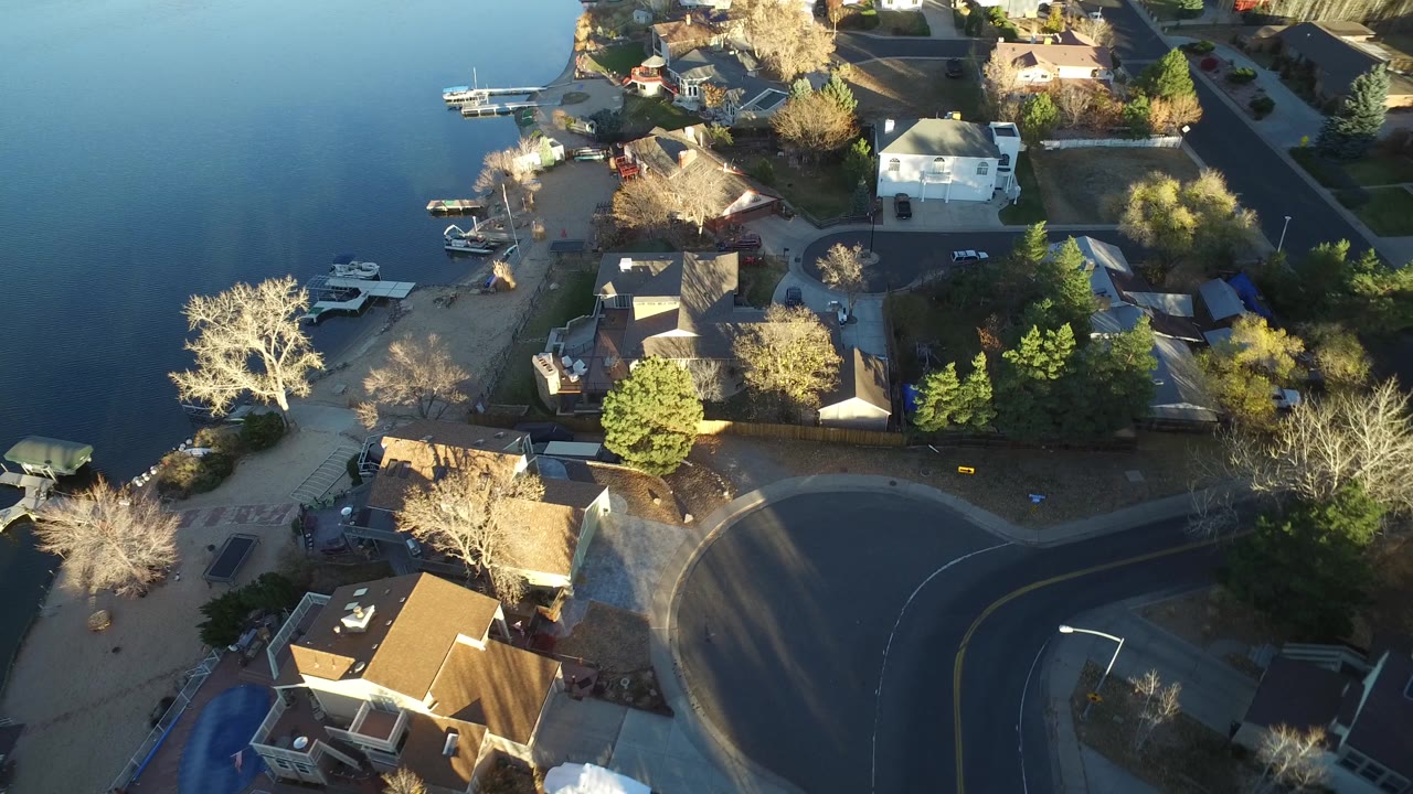Westminster, Colorado aerial frame with water on the left, docks and small boats near shore, houses in the center, and an empty curved road below, with no visible people.