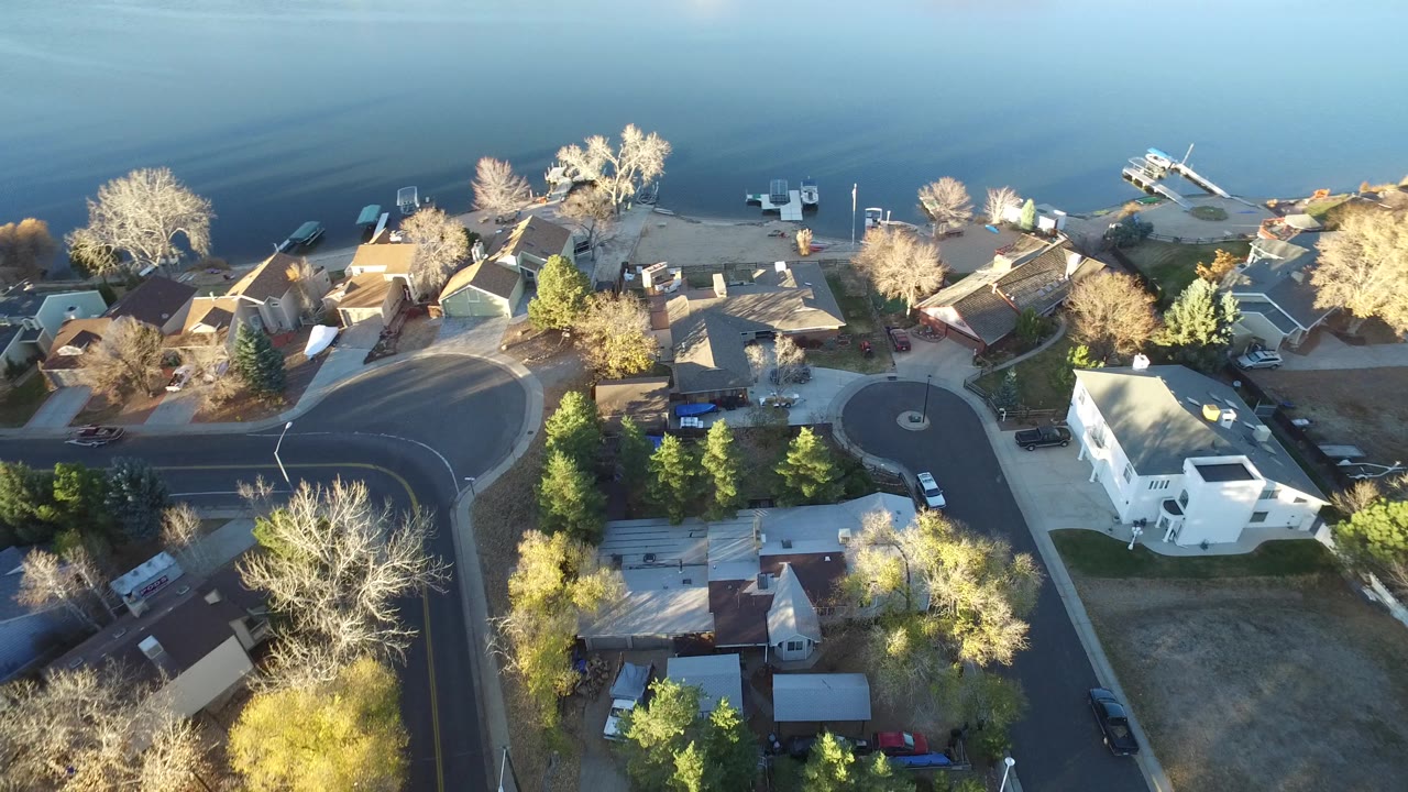 High overhead view in Westminster, Colorado with rooftops, a broad curved street, sunlit trees, lakeside docks, small boats, parked vehicles, and no visible people.