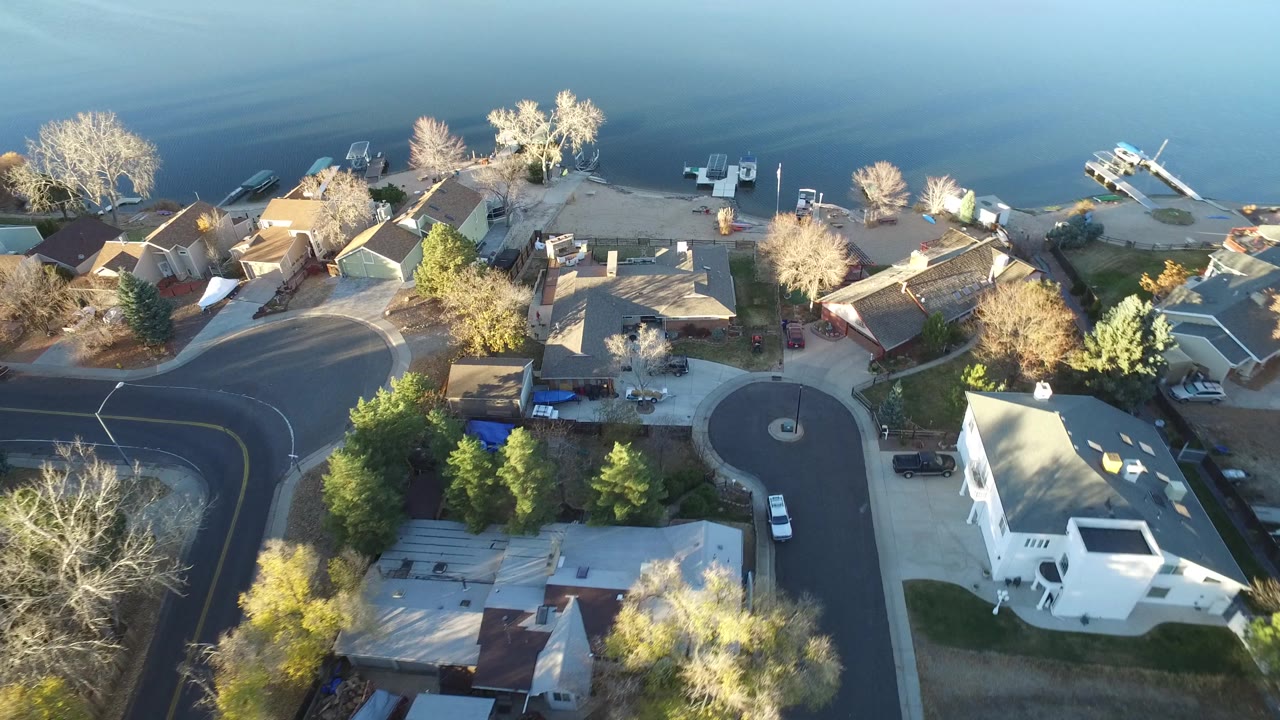 Elevated frame of Westminster, Colorado rooftops, a circular street end, mature trees, shoreline structures, and blue water, with no visible people.