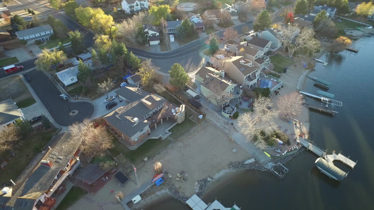 Overhead Westminster, Colorado frame with calm lake water at right, sandy beach below the houses, several docks, mixed trees, and no visible people.