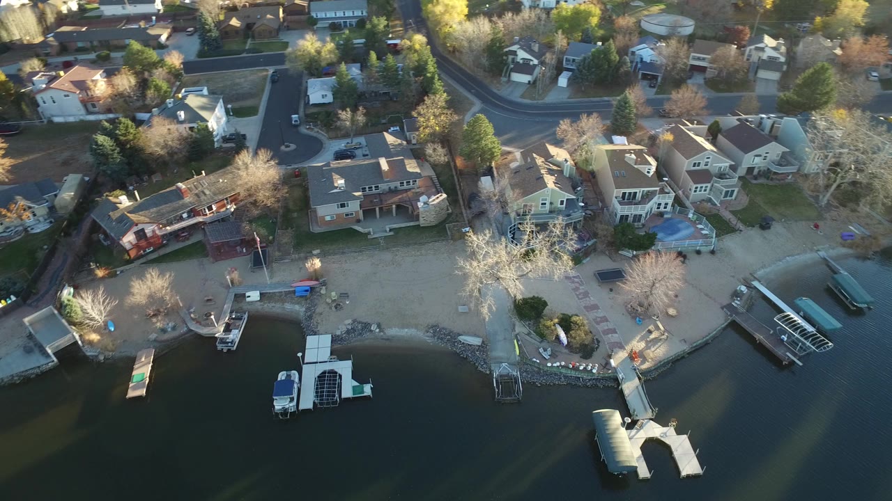 Aerial frame over Westminster, Colorado homes beside dark water, with docks, boats, bare trees, rooftops, and no visible people.