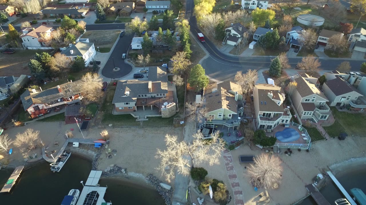 Aerial view in Westminster, Colorado showing residential roofs, leafless trees, parked vehicles, a curved road, sandy yards, docks, and no visible people discernible.