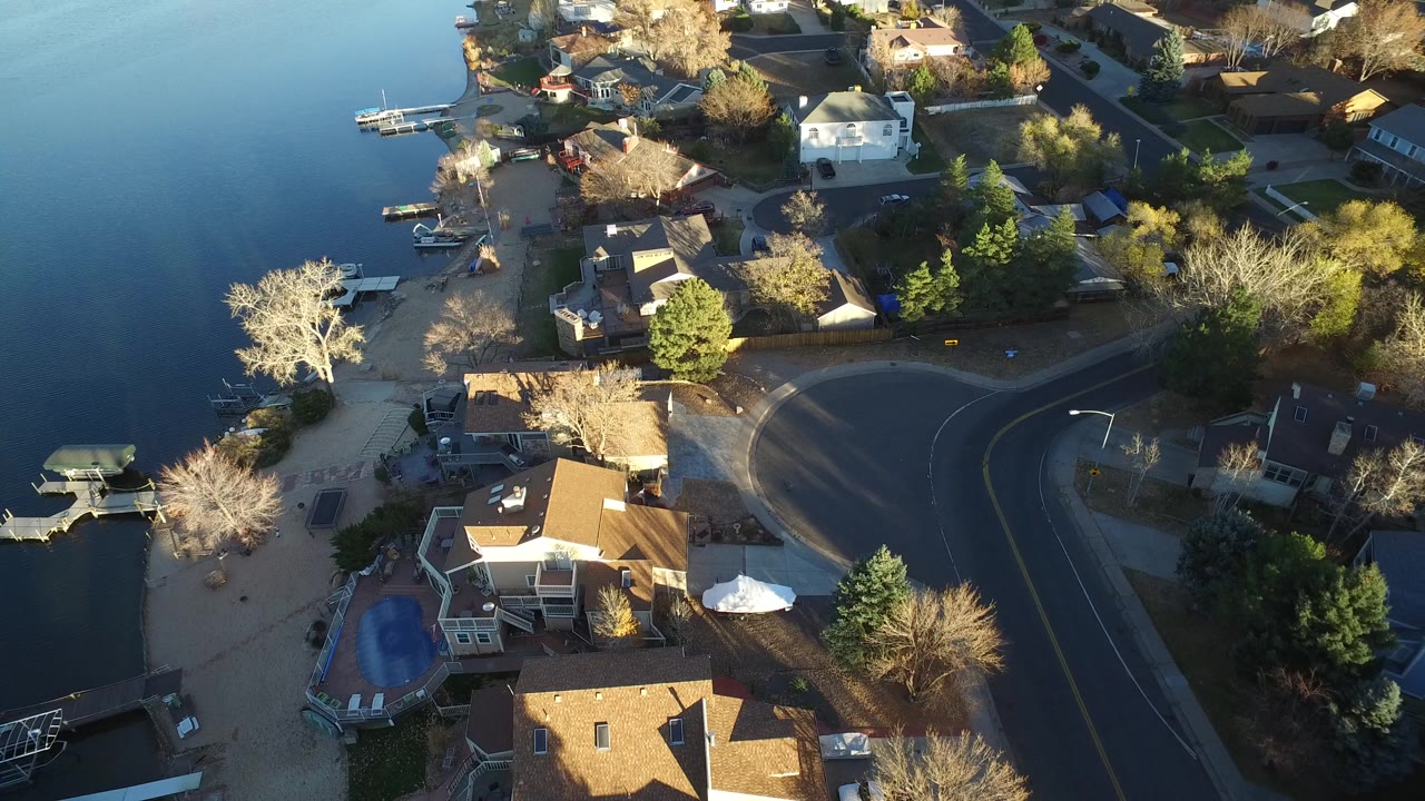 Aerial frame over Westminster, Colorado showing lake water, shoreline homes, small docks, boats, leafless trees, a curved road, and no visible people.