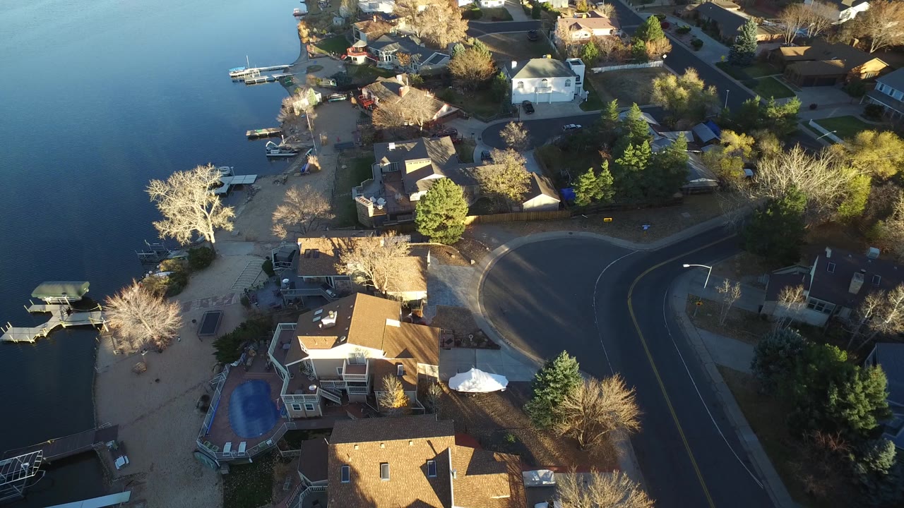 Drone-like overhead view of a lakeside block with houses, docks, boats, and trees.
