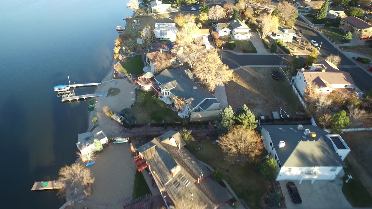 Aerial frame over Westminster, Colorado houses along the lake, with a dock and boats near the lower left, roofs and yards in warm light, and no visible people.