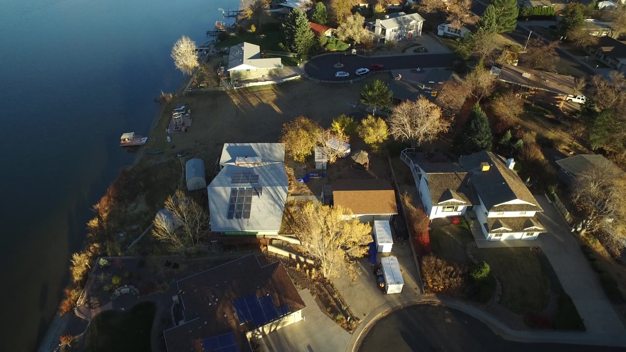 Drone view of homes near water with lawns, trees, parked cars, and residential roads.