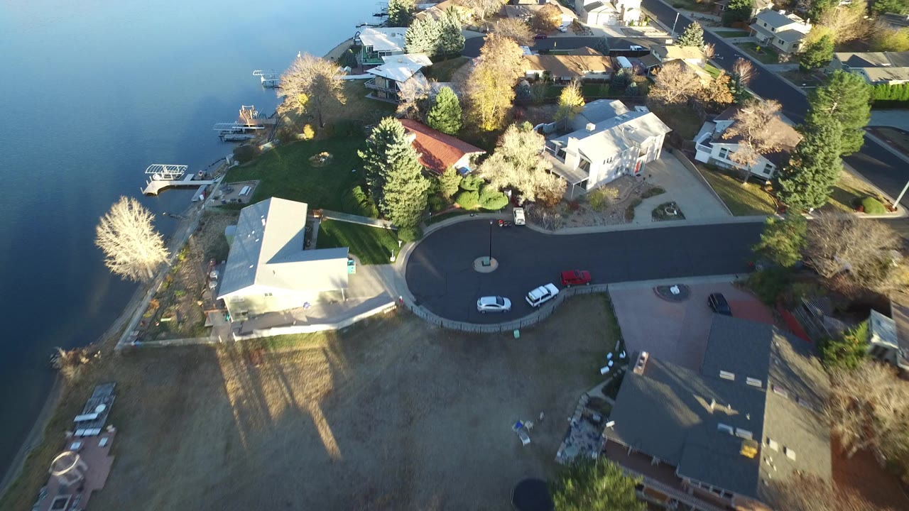 Aerial view over Westminster, Colorado lakefront homes, a curved residential street, parked cars, lawns, docks, and calm water at left, with no visible people.