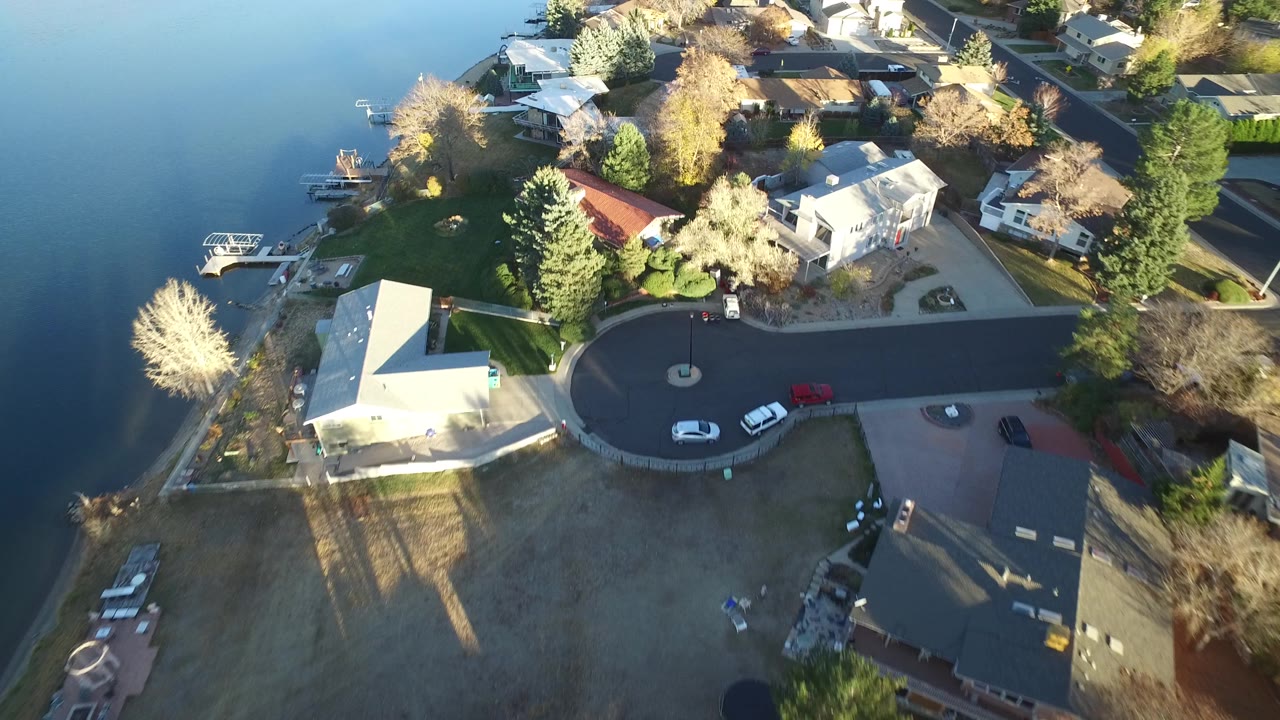 High aerial frame in Westminster, Colorado showing the lake edge on the left, a white house near a cul-de-sac, neighboring rooftops, parked vehicles, and no visible people.