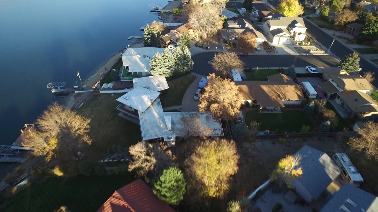 Overhead aerial frame in Westminster, Colorado showing calm lake water at left, private homes, yards, docks, parked vehicles, and autumn-colored trees, with no visible people.